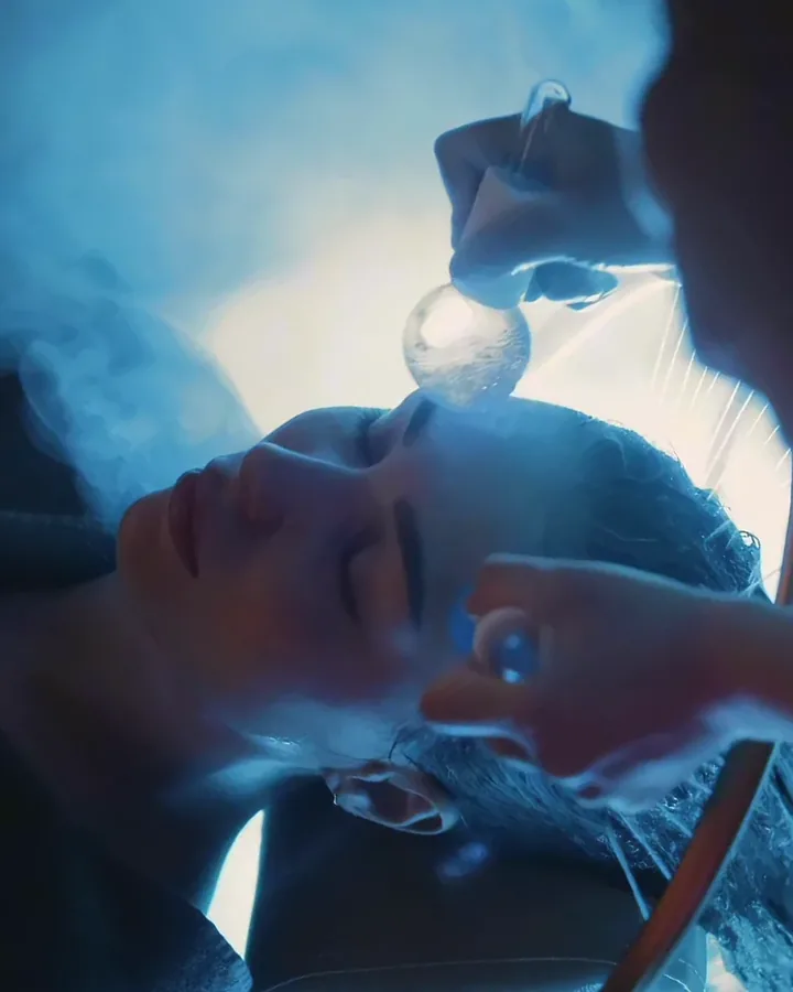A guest resting at the basin as glass globes are used during facial care, with mist under soft blue light
